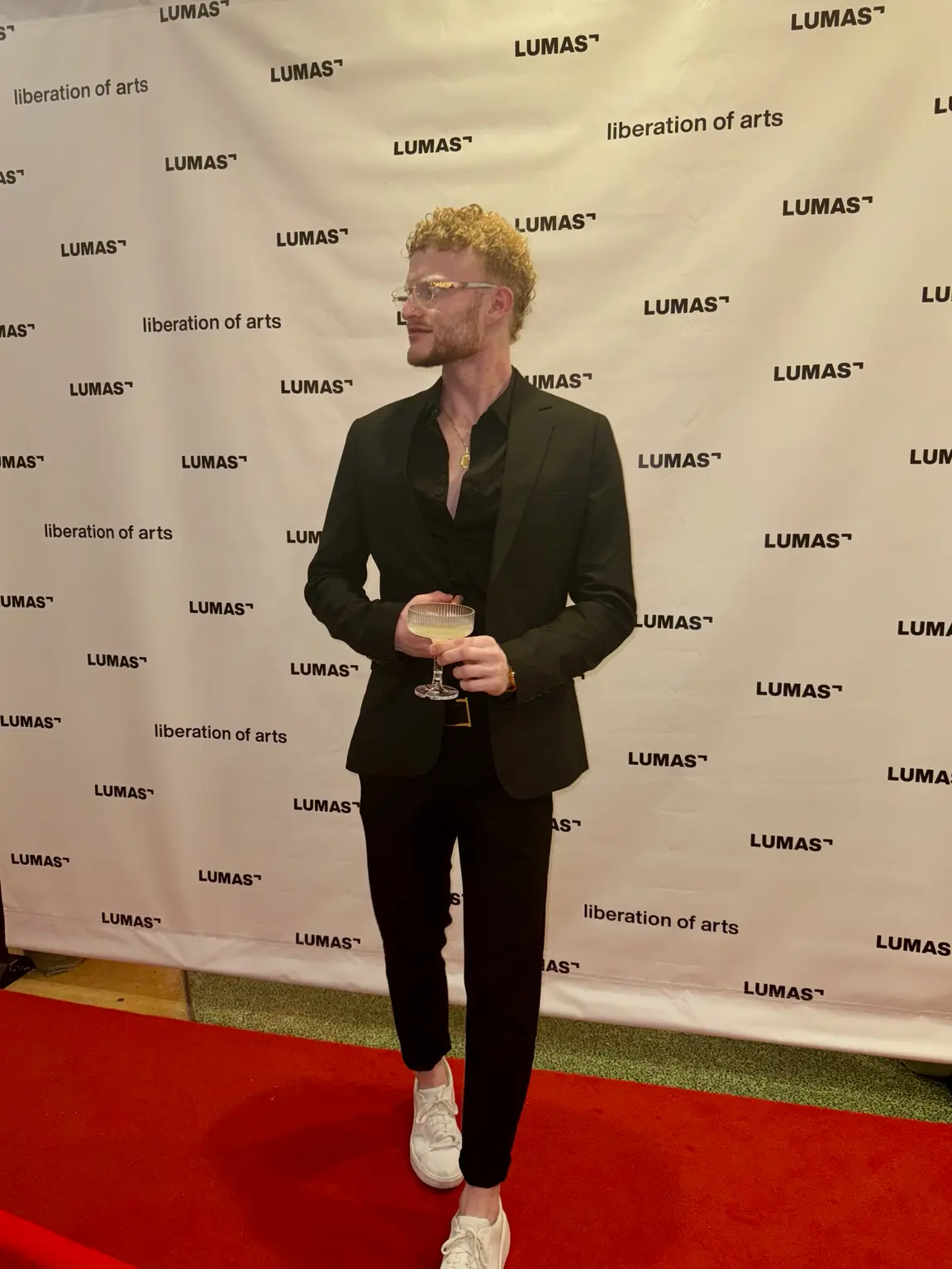 Royed in a black suit and black shirt, light sneakers and glasses, holding a glass on the red carpet before the LUMAS liberation of arts backdrop